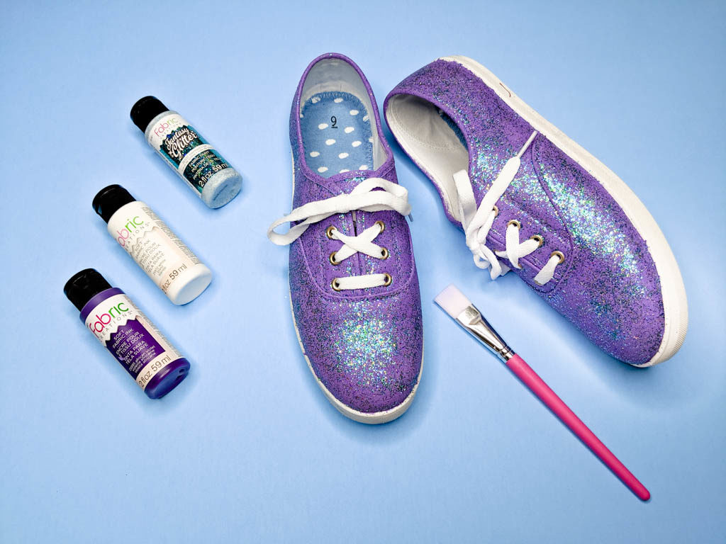 Purple and green glitter shoes set next to three paint bottles and a paint brush.