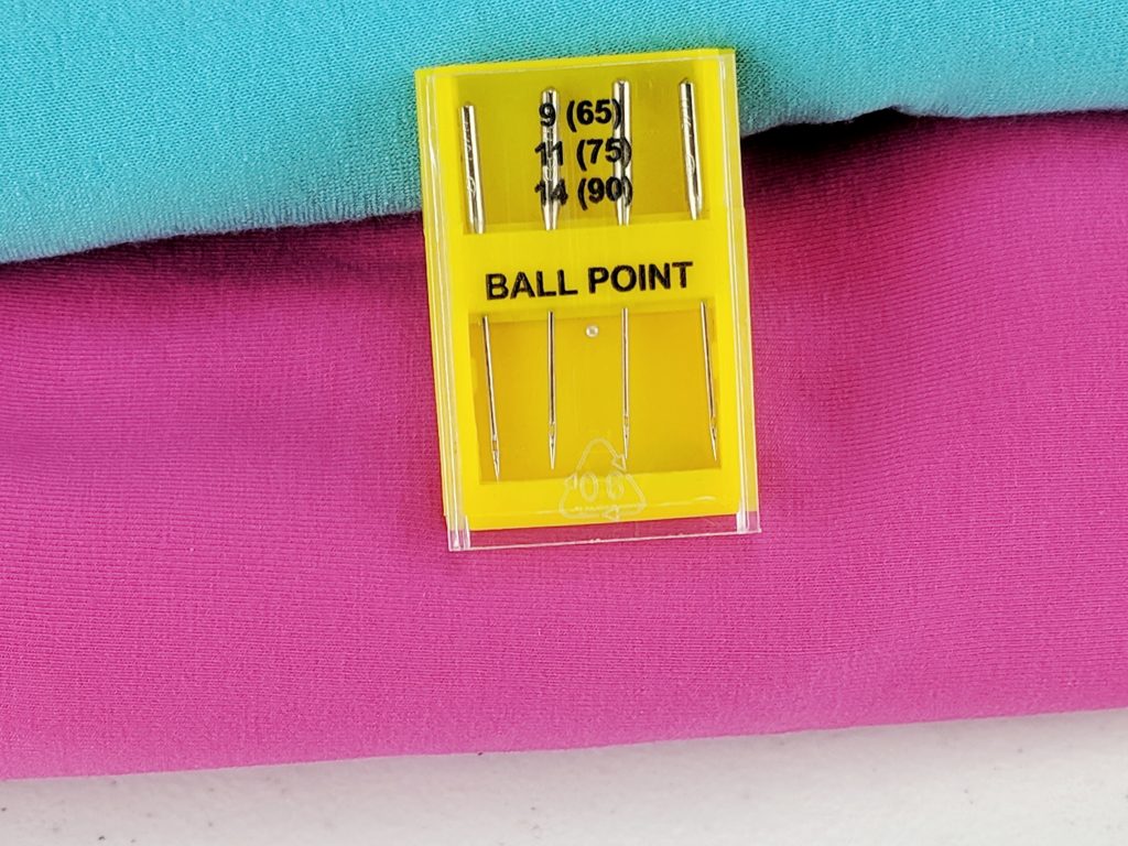 package of ball point needles laid on pink and blue fabric.