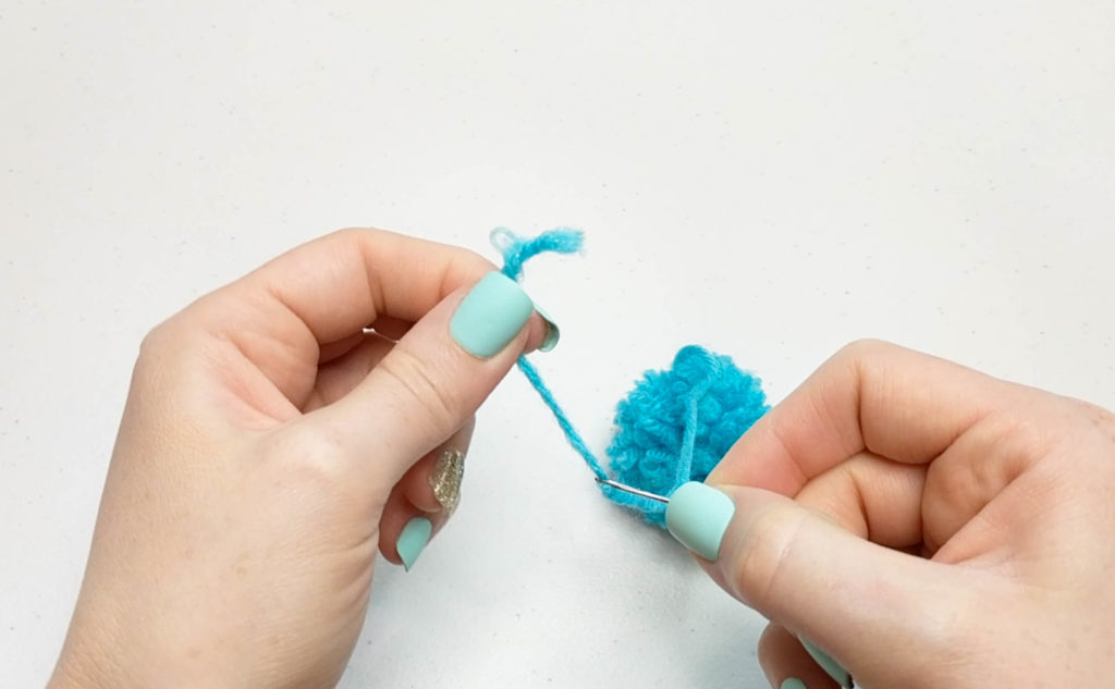 Embroidery needle being strung onto a pom pom with one long tail.