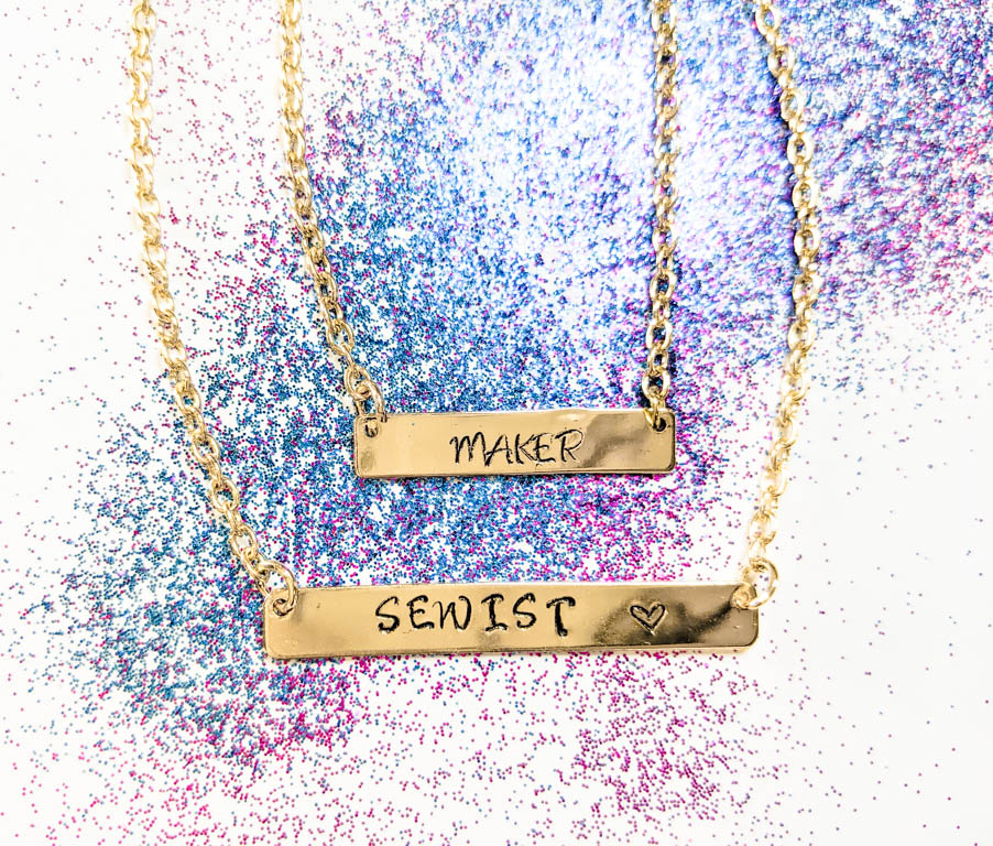 Two gold necklaces with the words "maker" and "sewist" stamped into them laying on top of blue and purple glitter.