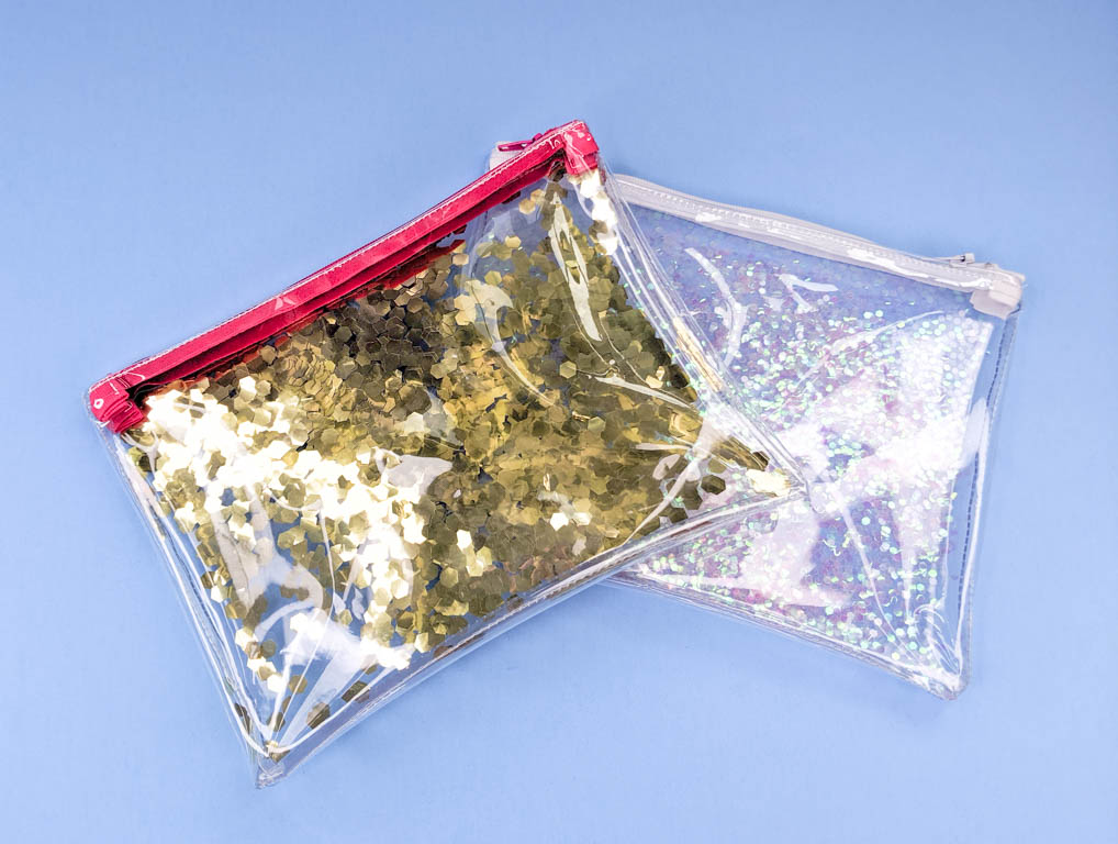 Two zipper bags made from clear vinyl and glitter. One has a pink zipper and gold glitter, the other has a white zipper and pink glitter. Showcased against a blue background