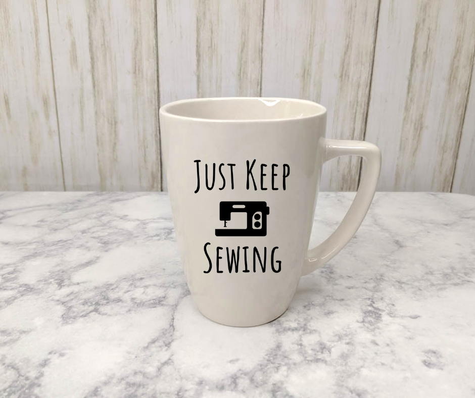 White mug with text "Just keep sewing" and an image of a sewing machine.