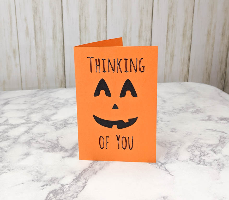 Orange paper card with a jack-o-lantern face and the text "thinking of you" written on it.
