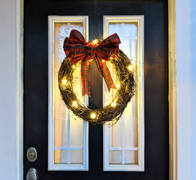 Christmas light wreath made with grapevine and with lights shaped as stars and a big plaid red bow. Lights are turned on