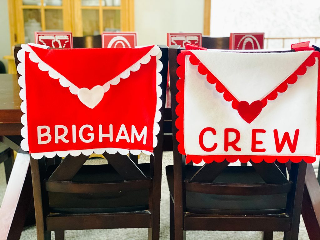 Two heart chair covers on chairs. One is red with white trim and the other is opposite in colors. They are made to look like envelopes with names on them.