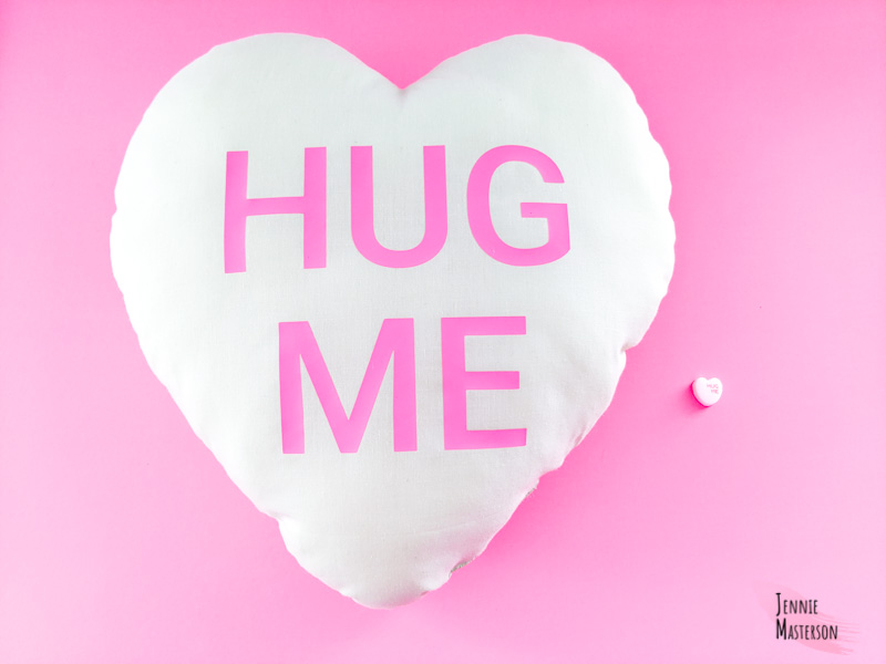 Conversation hart pillow next to a conversation heart candy. both are white and say "hug me".