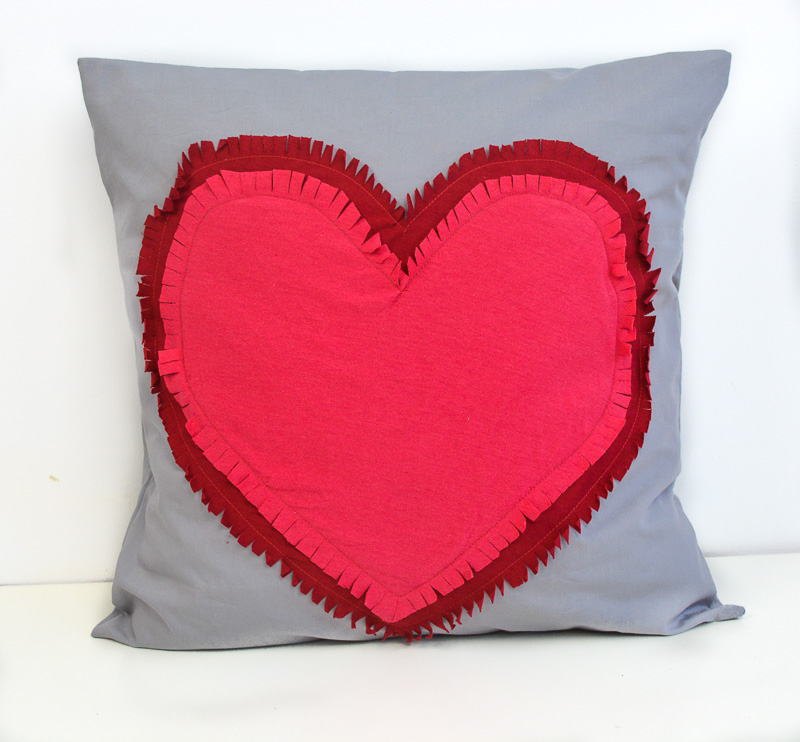 Gray square pillow with two hearts sewn on top of each other in the gray pillow center. The hearts have fringe.
