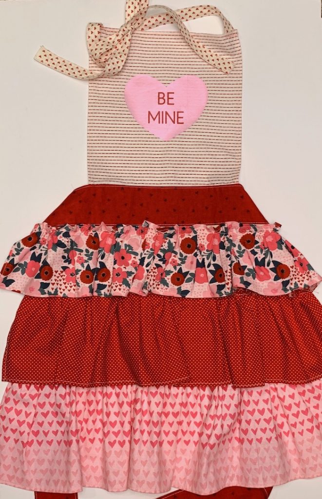 Valentine themed apron with gathered frills on the skirt. Top center features a heart that says "be mine".