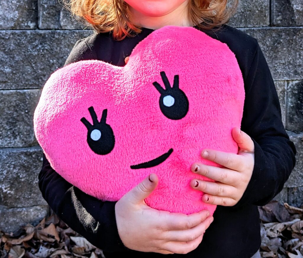 Hot pink heart plushie with face and eye lashes being held by a little girl.